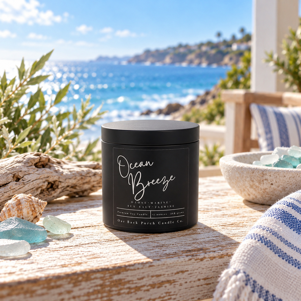 Ocean Breeze matte black tin soy candle by Back Porch Candle Co. on a coastal table with sea glass, seashells, driftwood, striped linen, and ocean views.