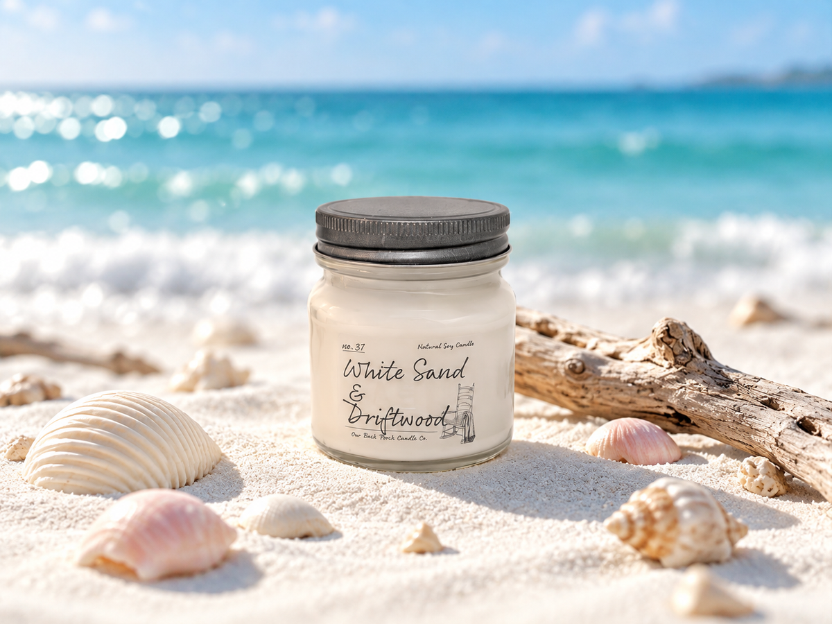 Back Porch Candle Co White Sand & Driftwood 8 oz soy candle on white sand with seashells and driftwood in a bright coastal setting.
