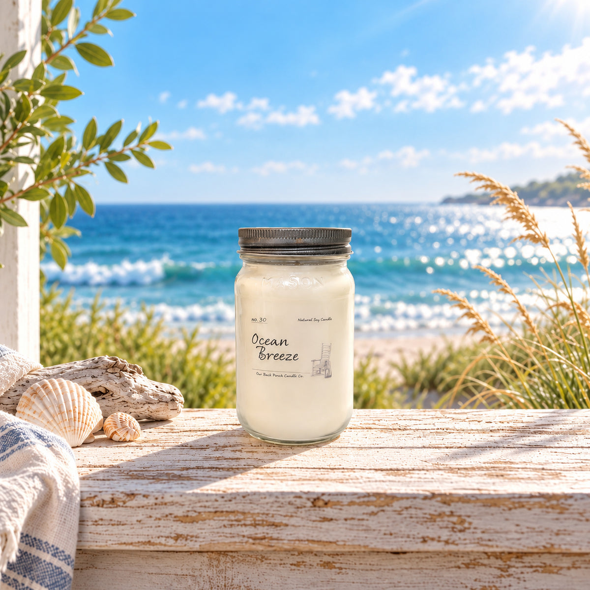Ocean Breeze 16 oz soy candle by Back Porch Candle Co. in a bright coastal setting with sea, shells, driftwood, and soft natural light.